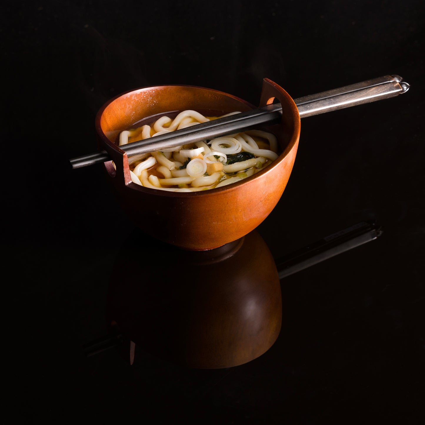 A pair of black wooden chopsticks with a carved lotus flower tip sitting accross the top of a noodle bowl filled with noodles.